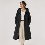 A woman modeling a long black hooded coat over a white button-up shirt and beige trousers, standing against a plain light gray studio background.