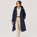 A woman modeling a navy blue hooded raincoat over a cream ribbed sweater, cream trousers, and a brown belt, photographed against a clean light gray studio background.