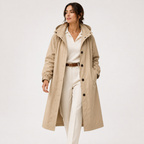 A woman modeling a mid-length beige hooded trench coat over a white collared shirt and white trousers, photographed against a clean light gray studio background.