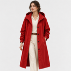A woman modeling a long red hooded trench coat over a cream collared shirt, cream trousers, and a brown belt, photographed against a clean light gray studio background.