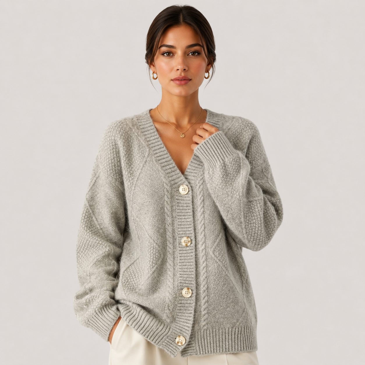 A woman wearing a light gray cable-knit cardigan sweater with cream buttons, paired with off-white trousers, against a solid light gray studio background.