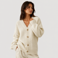 A woman wearing a cream-colored cable-knit cardigan sweater with brown buttons, paired with matching cream trousers, set against a solid light gray background.