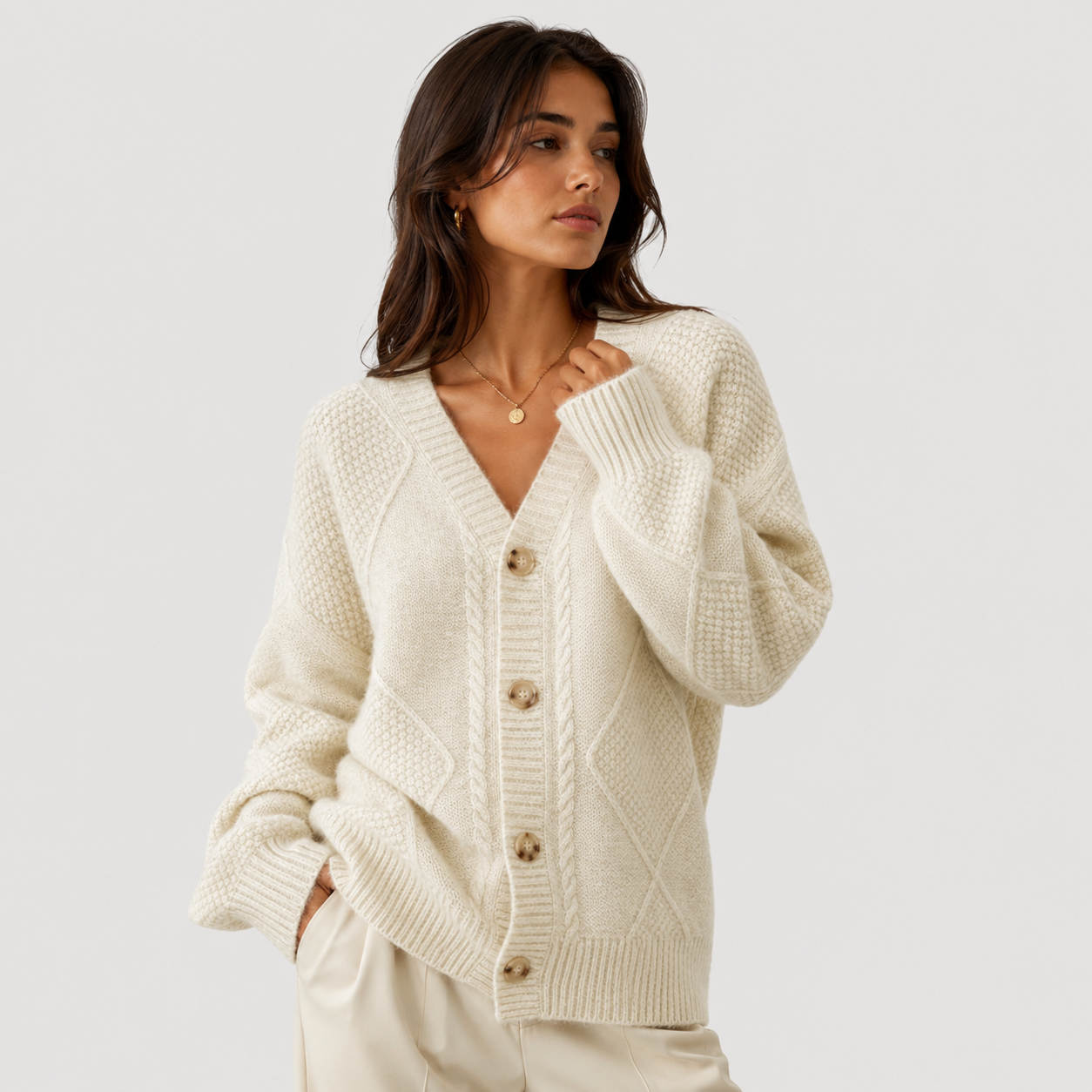 A woman wearing a cream-colored cable-knit cardigan sweater with brown buttons, paired with matching cream trousers, set against a solid light gray background.