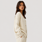A woman wearing a cream-colored, textured cable-knit cardigan sweater with buttons, paired with matching cream trousers, photographed against a solid light gray studio background.
