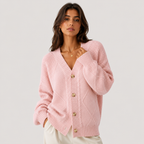 A woman wearing a light pink cable-knit cardigan sweater with decorative wooden buttons, paired with white pants, photographed against a solid light gray studio background.