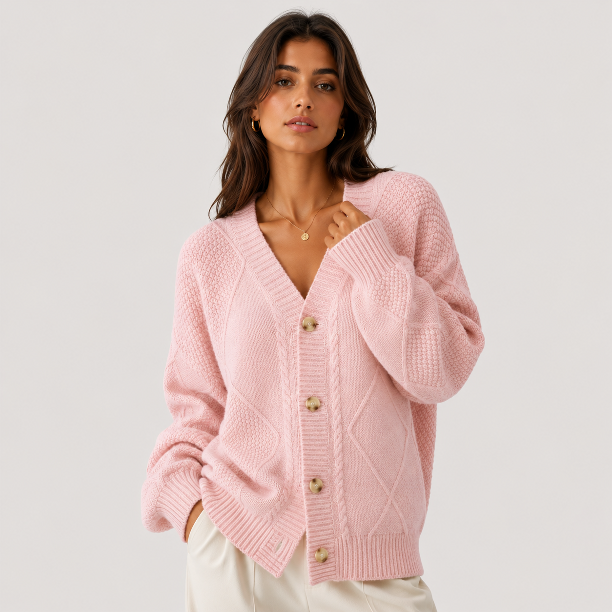 A woman wearing a light pink cable-knit cardigan sweater with decorative wooden buttons, paired with white pants, photographed against a solid light gray studio background.