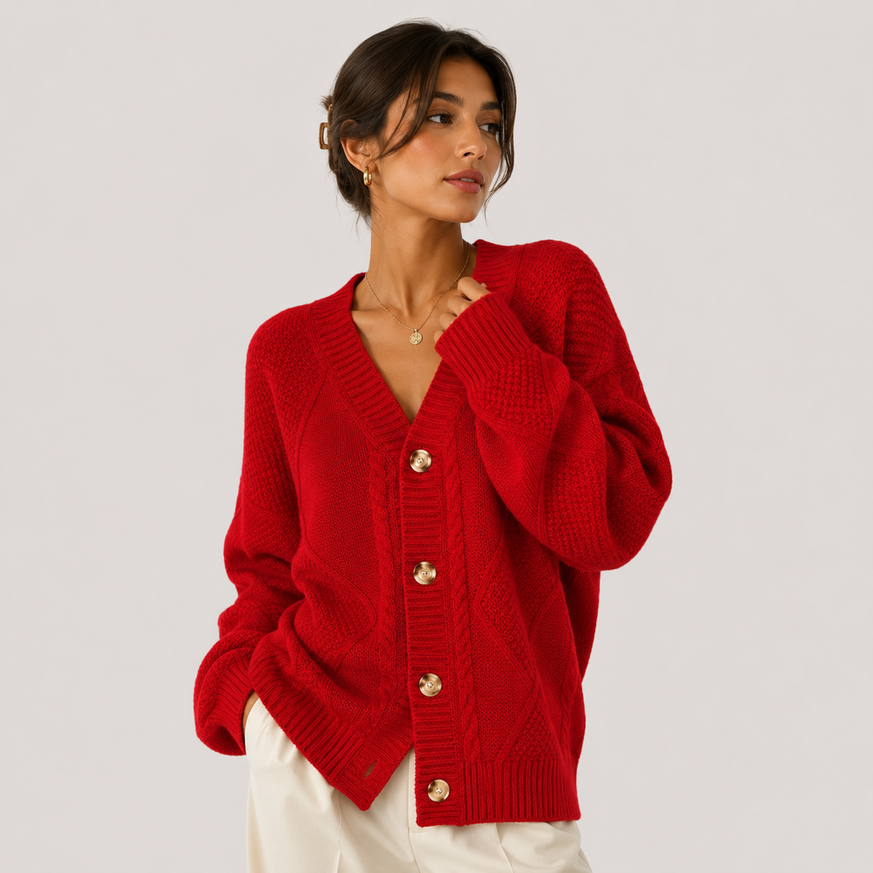 A woman wearing a bright red cable-knit cardigan sweater with gold-tone buttons, styled with white pants against a light gray studio background.