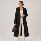 A woman models a long black trench coat over a cream top and cream wide-leg trousers, holding a dark brown structured handbag against a plain light gray studio background.