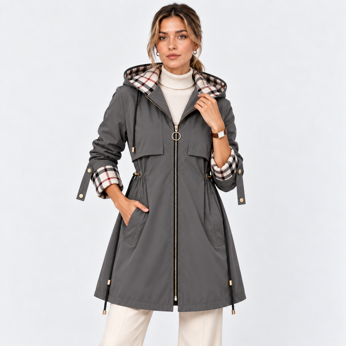 A woman models a dark gray hooded coat with a beige, brown, and black plaid lining on the hood and cuffs, paired with a white turtleneck and cream pants, against a light gray studio background.