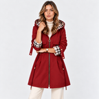 A woman modeling a red hooded parka jacket with a black, white, and red plaid lining on the hood and cuffs, paired with a white turtleneck and cream pants, against a light gray studio background.