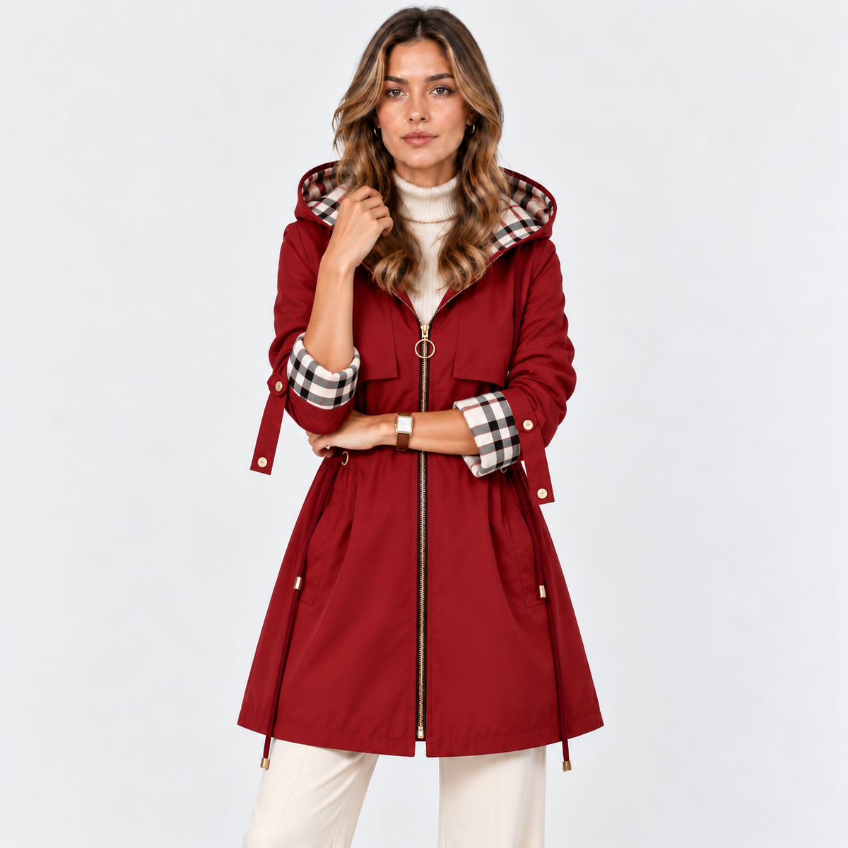 A woman modeling a red hooded parka jacket with a black, white, and red plaid lining on the hood and cuffs, paired with a white turtleneck and cream pants, against a light gray studio background.