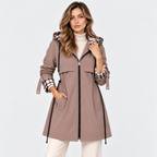 A woman models a taupe-brown hooded raincoat with a black front zipper, black drawstrings, and a plaid pattern lining the hood and cuffs, paired with a white turtleneck and white pants against a light gray studio background.