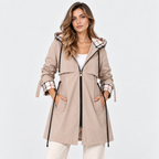 A woman modeling a mid-length beige hooded parka jacket with a black front zipper, black drawstrings, and plaid trim on the hood and cuffs, paired with white pants against a light gray studio background.