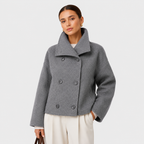 A woman modeling a gray double-breasted cropped coat with a wide collar, paired with cream trousers, against a light gray studio background.