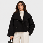 A woman models a black cropped double-breasted coat with a wide lapel over a cream turtleneck and cream trousers, holding a black handbag against a light gray studio background.