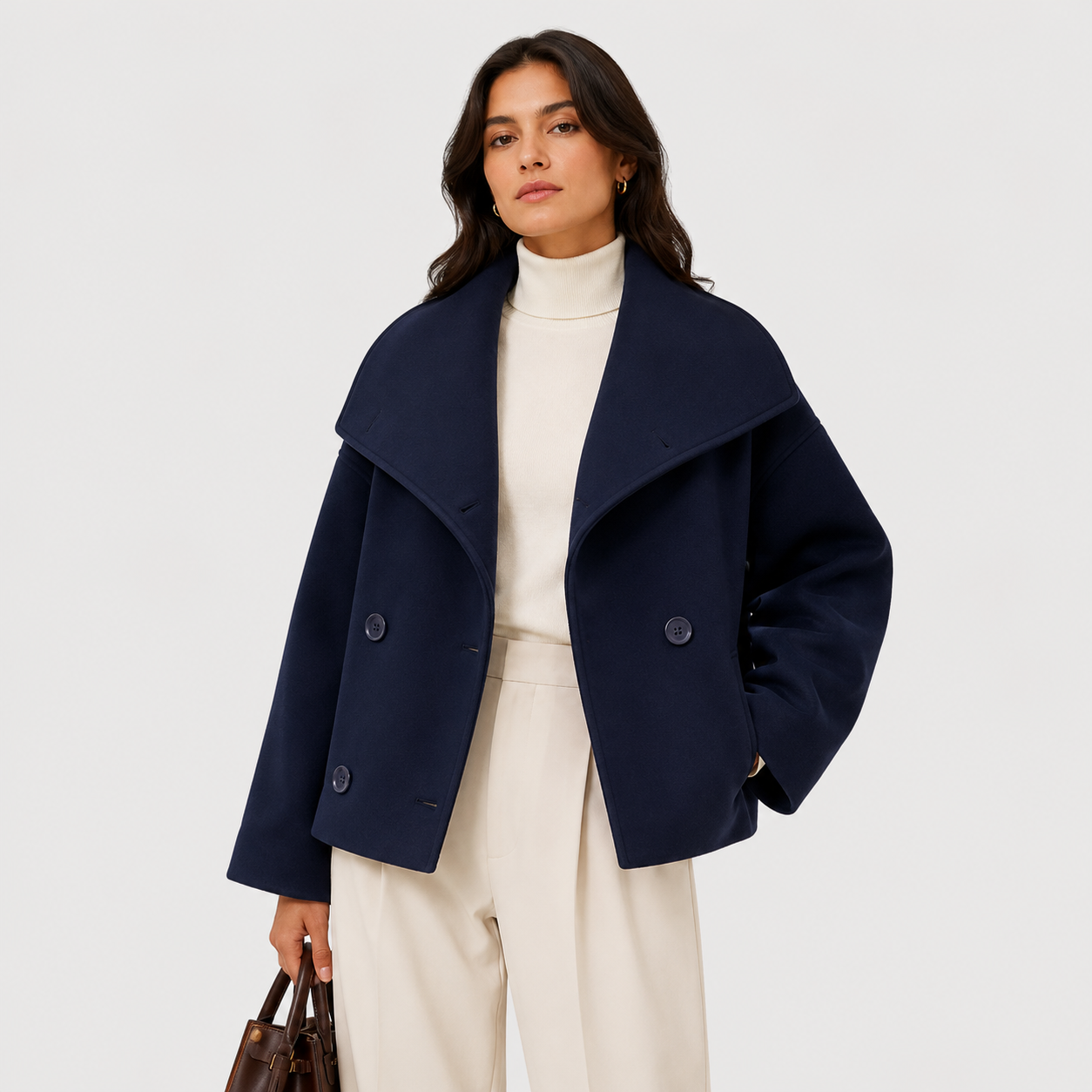 A woman modeling a navy blue double-breasted coat with a wide shawl collar, worn over a cream turtleneck and cream wide-leg trousers, holding a brown handbag against a light gray studio background.
