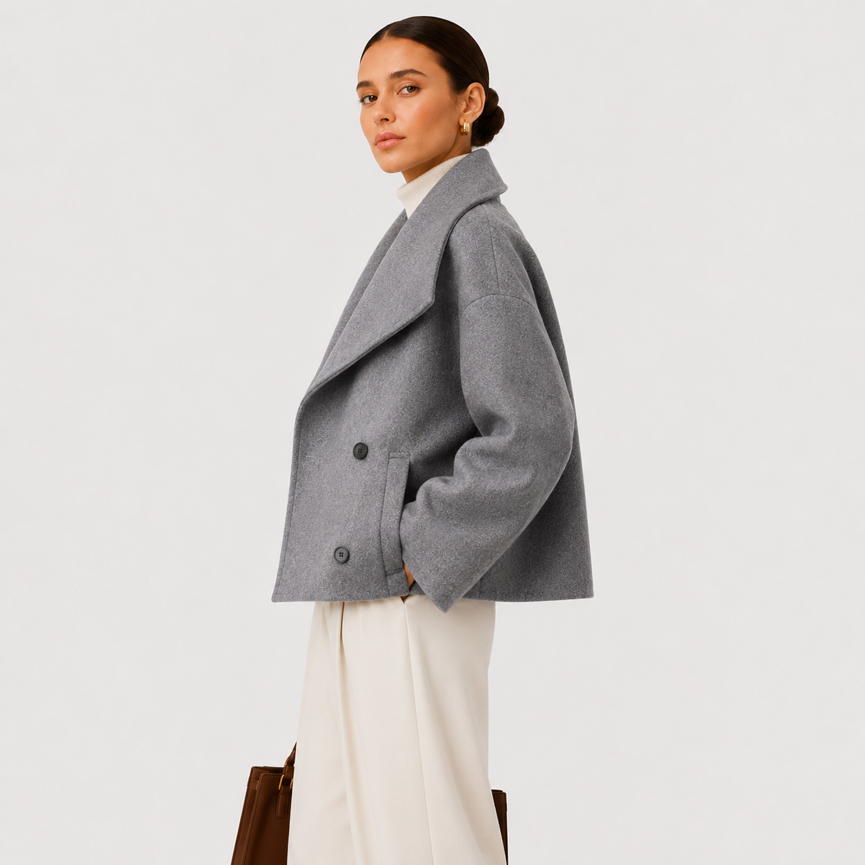 A woman modeling a gray double-breasted wrap-style coat over a white top and cream wide-leg trousers, standing against a plain light gray studio background.