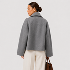 A woman shown from the back wearing a cropped gray wool-blend jacket with a high collar, paired with cream wide-leg trousers and holding a brown leather handbag, against a light gray studio background.