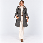 A woman models a dark gray trench coat with a beige and brown plaid lining on the collar and cuffs, worn over a cream turtleneck and cream wide-leg trousers, against a light gray studio background.