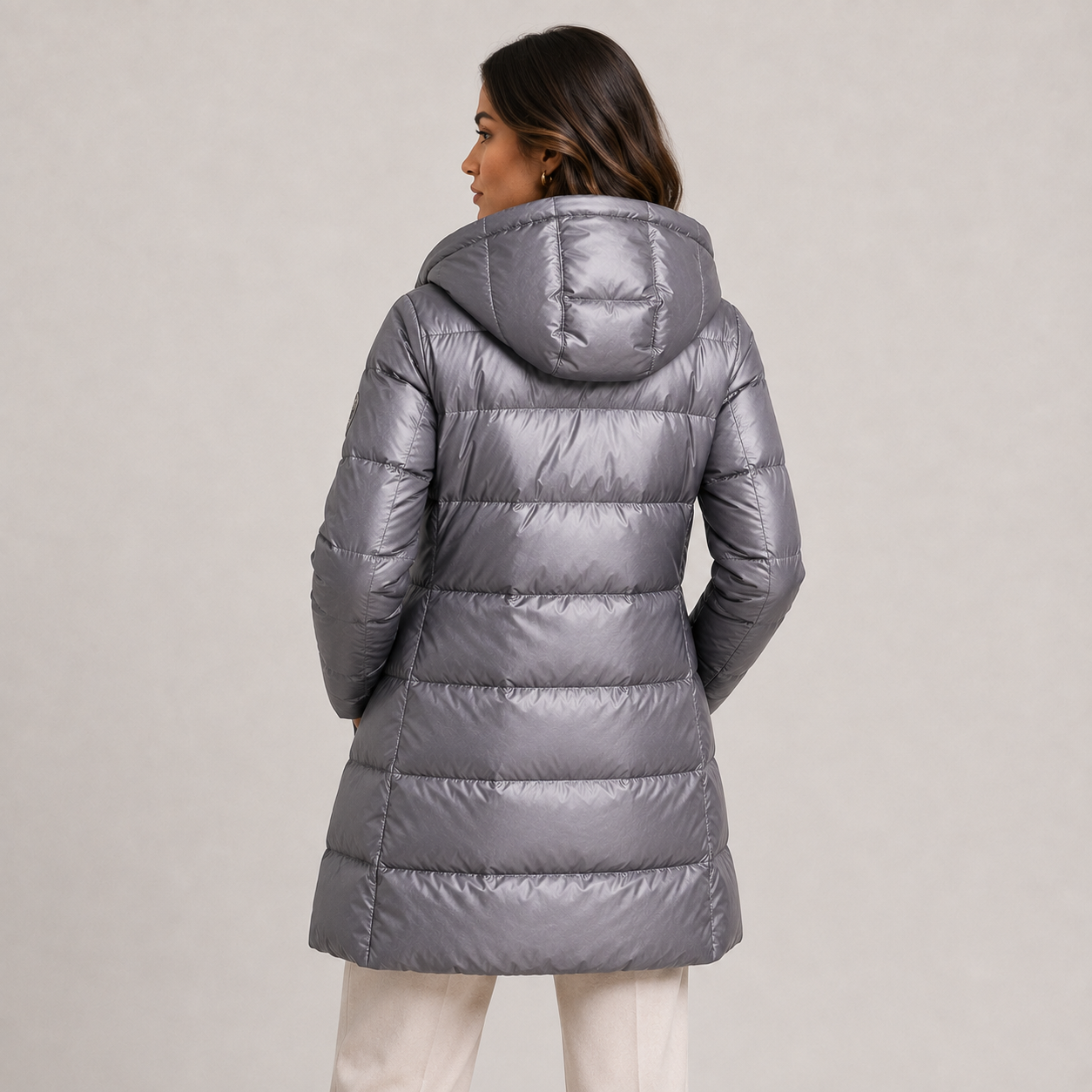 A woman shown from the back wearing a shiny silver-gray hooded puffer jacket, paired with off-white pants, against a plain light gray studio background.