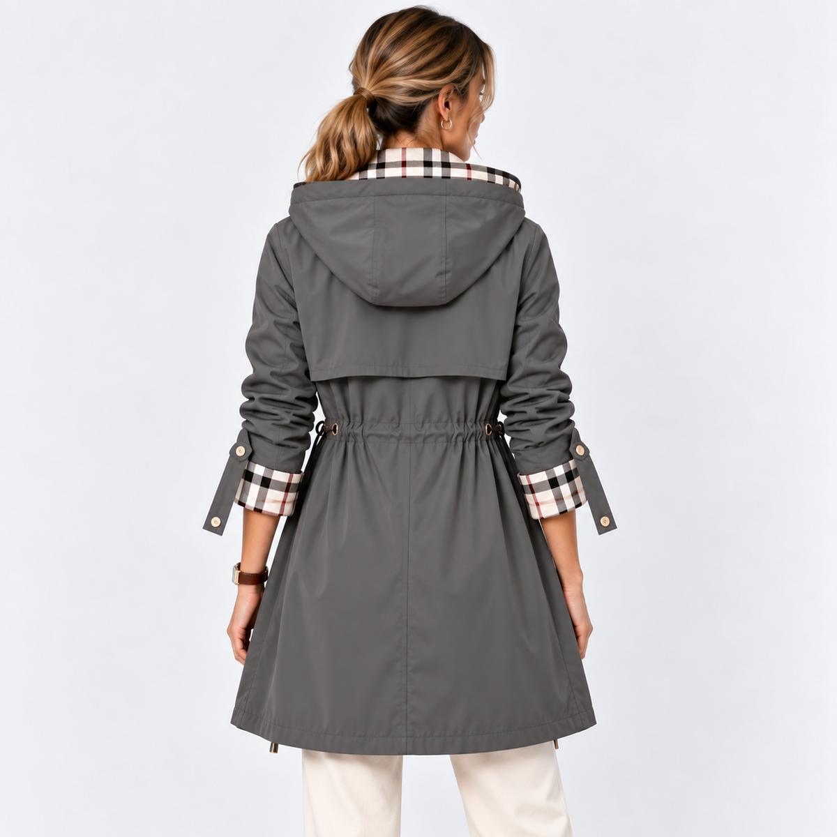 A woman viewed from the back wearing a dark gray hooded trench coat with a beige, black, and white check patterned collar lining and matching checkered cuffs, paired with off-white pants against a light gray studio background.