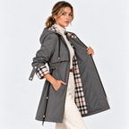 A woman models a gray hooded raincoat with a beige, black, and white plaid lining. She is wearing a white turtleneck and white trousers, posing against a light gray studio background.