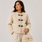 A woman models a cream-colored sherpa fleece cropped jacket with black toggle buttons, paired with cream trousers and holding a matching cream handbag, set against a plain light gray studio background.