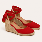 A pair of red wedge sandals with espadrille-style jute-wrapped wedge heels and adjustable ankle straps tied in bows, shown on a light beige background.