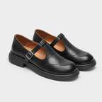 A pair of black leather Mary Jane shoes with brogue detailing and adjustable straps, displayed on a light gray background.