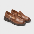 A pair of women's Mary Jane wingtip shoes in brown leather, featuring a strap with a silver buckle, decorative brogue detailing, and low stacked heels, shown on a light gray background.