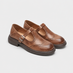 A pair of women's Mary Jane wingtip shoes in brown leather, featuring a strap with a silver buckle, decorative brogue detailing, and low stacked heels, shown on a light gray background.