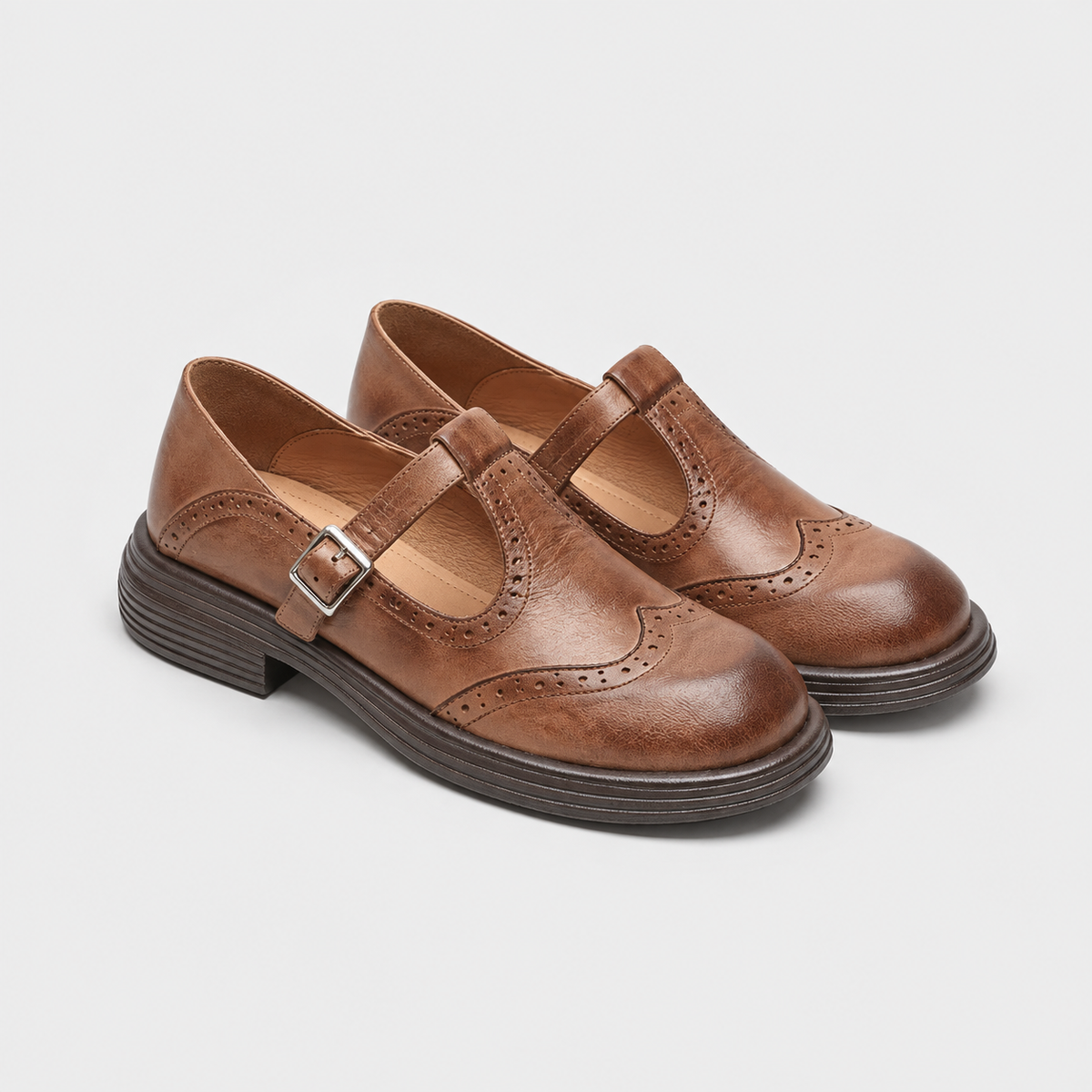 A pair of women's Mary Jane wingtip shoes in brown leather, featuring a strap with a silver buckle, decorative brogue detailing, and low stacked heels, shown on a light gray background.