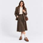 A woman models a brown plaid skirt suit consisting of a blazer and a pleated midi skirt, styled with a cream knit shirt, brown leather shoes, white socks, and a brown handbag against a light gray studio background.