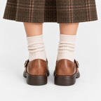 Rear view of a person wearing brown leather buckle-strap loafers with dark brown ridged soles, cream ribbed socks, and a brown plaid skirt against a light gray studio background.