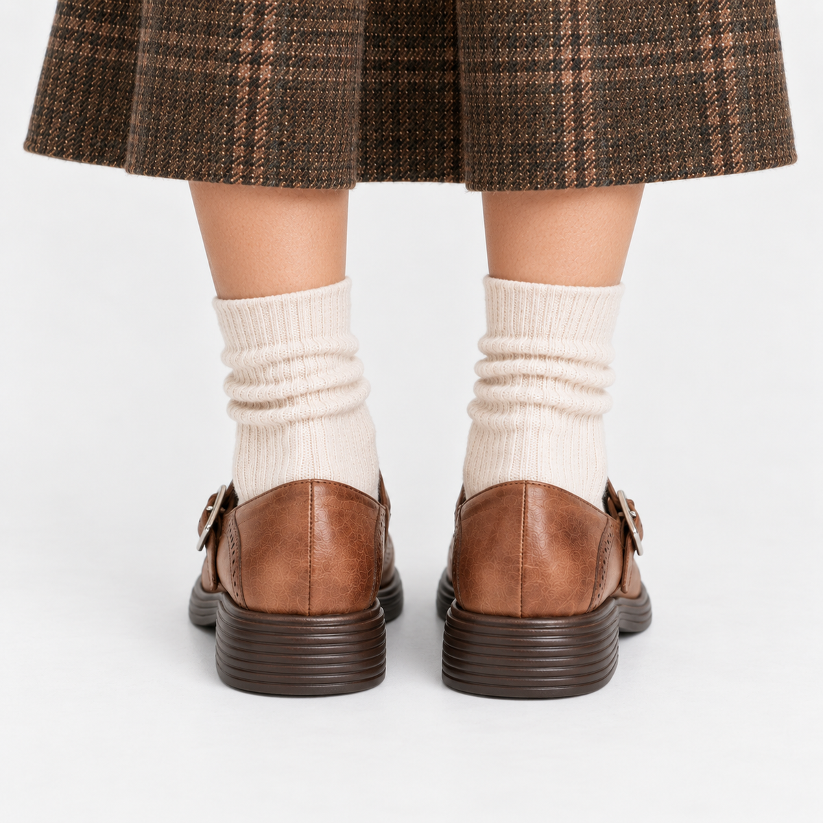 Rear view of a person wearing brown leather buckle-strap loafers with dark brown ridged soles, cream ribbed socks, and a brown plaid skirt against a light gray studio background.