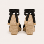A pair of women's espadrille wedge sandals shown from the back, featuring a woven jute platform, black fabric wedge inserts, and black ribbon ankle straps tied in bows with small beige tags.