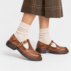 A pair of brown leather Mary Jane shoes with brogue detailing and a buckle strap, shown on a child's feet with cream ribbed socks and a brown plaid skirt against a light gray studio background.
