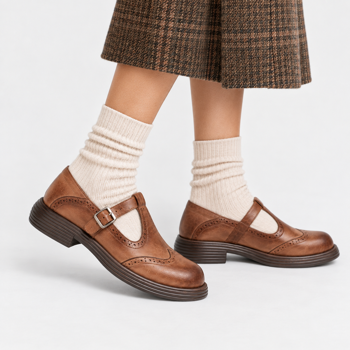 A pair of brown leather Mary Jane shoes with brogue detailing and a buckle strap, shown on a child's feet with cream ribbed socks and a brown plaid skirt against a light gray studio background.