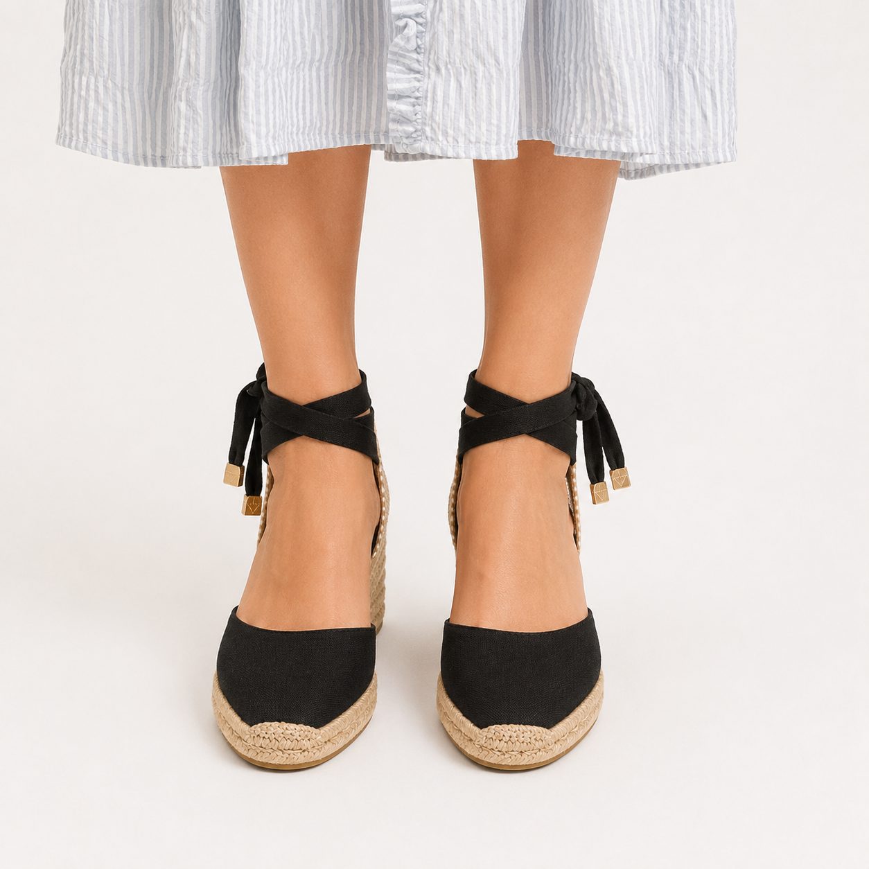 A pair of women's black espadrille wedge sandals with lace-up ankle straps and braided jute soles, shown on a model wearing a light blue and white striped skirt against a plain white background.