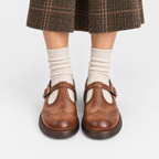 A pair of brown leather Mary Jane shoes with brogue detailing and buckle straps, worn with cream ribbed socks and a brown plaid skirt, shown from the knees down on a light gray background.
