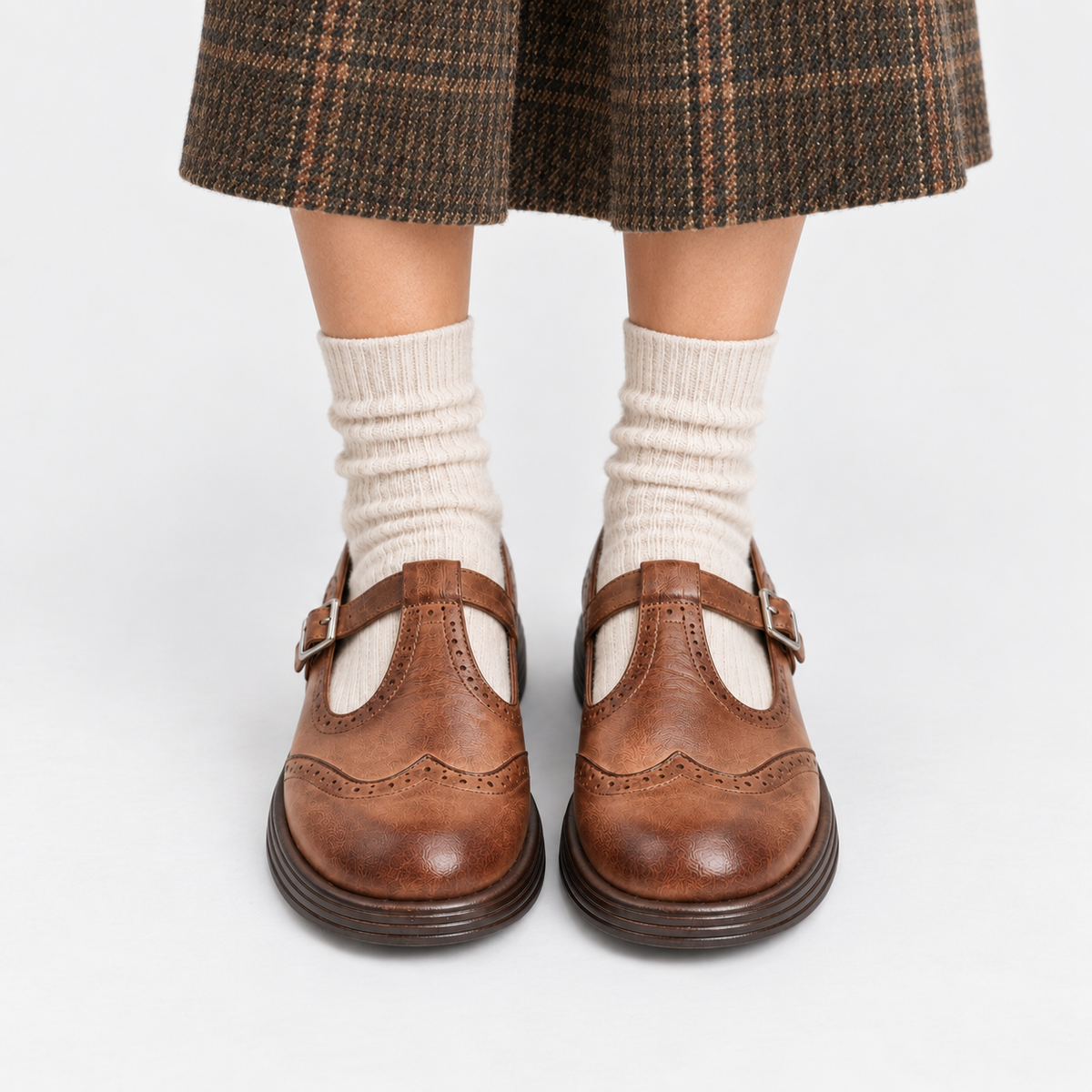 A pair of brown leather Mary Jane shoes with brogue detailing and buckle straps, worn with cream ribbed socks and a brown plaid skirt, shown from the knees down on a light gray background.