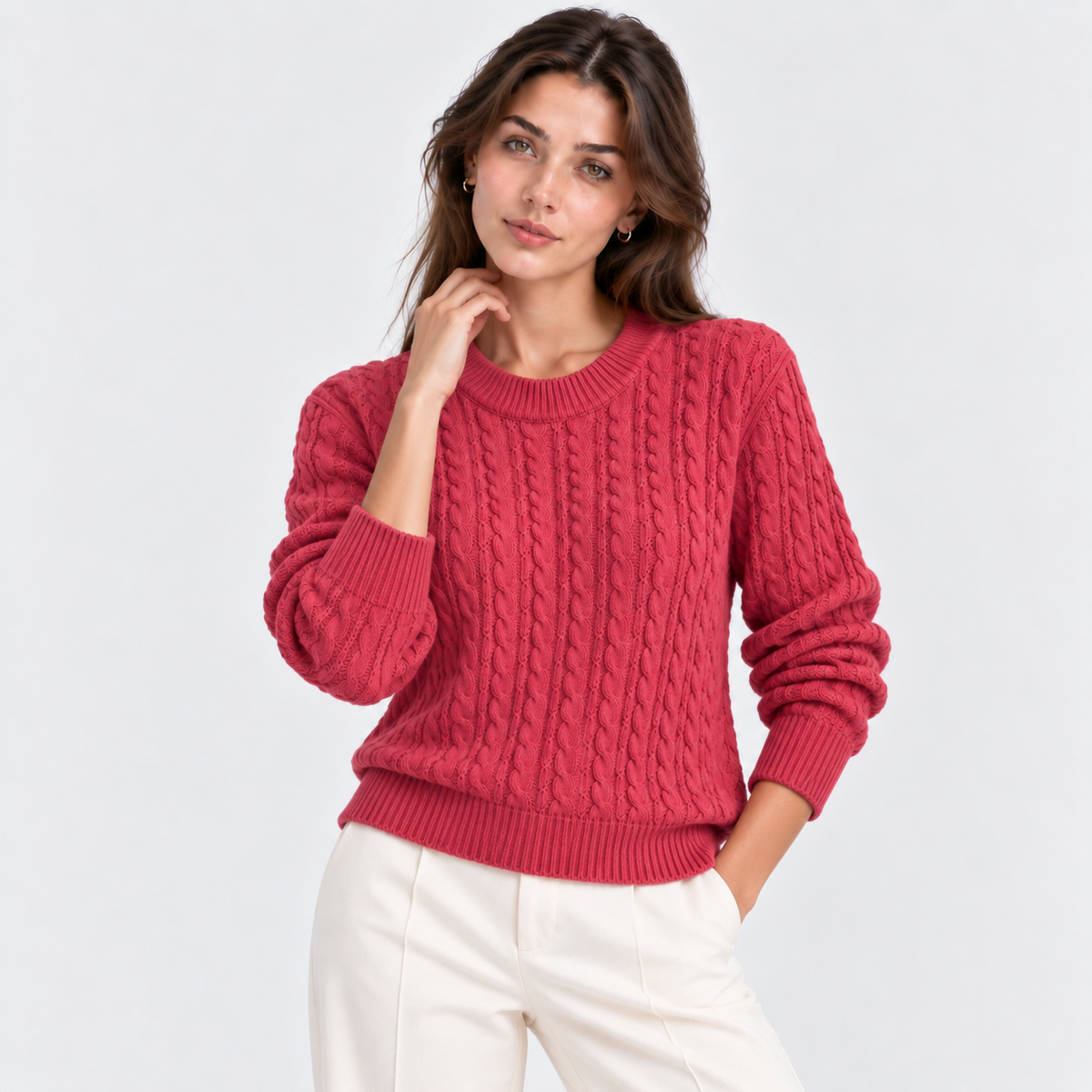 A woman wearing a vibrant red cable-knit crewneck sweater and white trousers, posing against a light gray studio background.