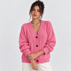 A woman wearing a pink chunky knit cardigan with dark buttons, paired with white pants, photographed against a light gray studio background.