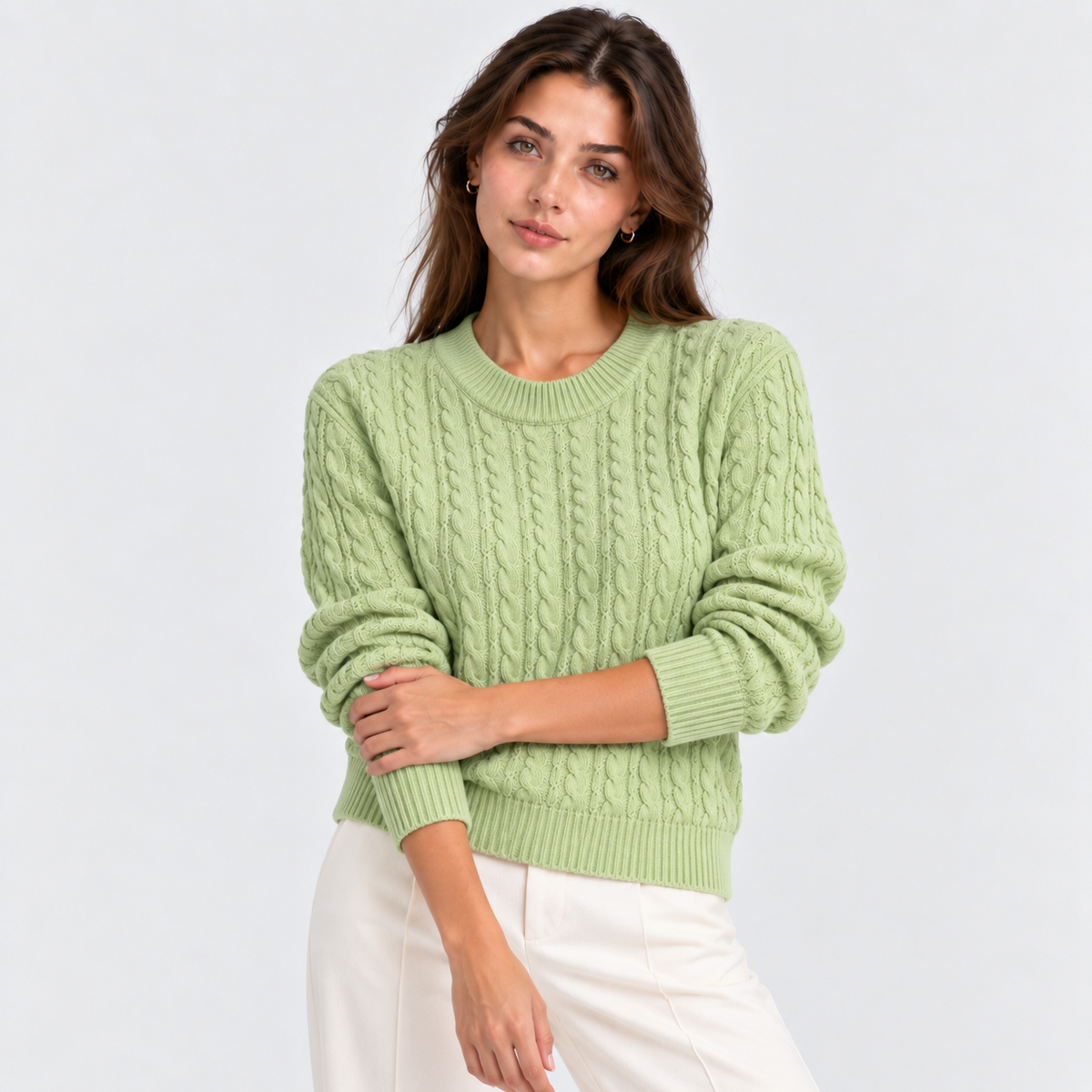 A woman wearing a light sage green cable-knit crewneck sweater and white pants, photographed against a clean light gray studio background.