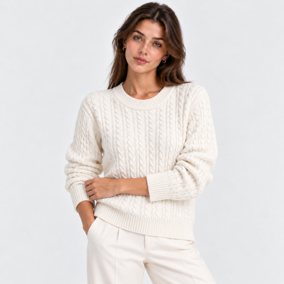 A woman wearing a cream cable-knit crewneck sweater and matching cream trousers, photographed against a clean light gray studio background.