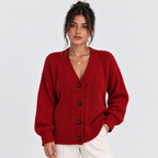 A woman wearing a red cable-knit cardigan sweater with dark buttons, paired with white pants, photographed against a light gray studio background.