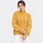 A woman wearing a mustard yellow cable-knit mock neck sweater and off-white trousers, standing against a light gray studio background.