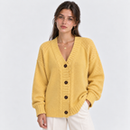 A woman wearing a mustard yellow cable-knit cardigan sweater with dark brown buttons, paired with white pants, against a light gray studio background.