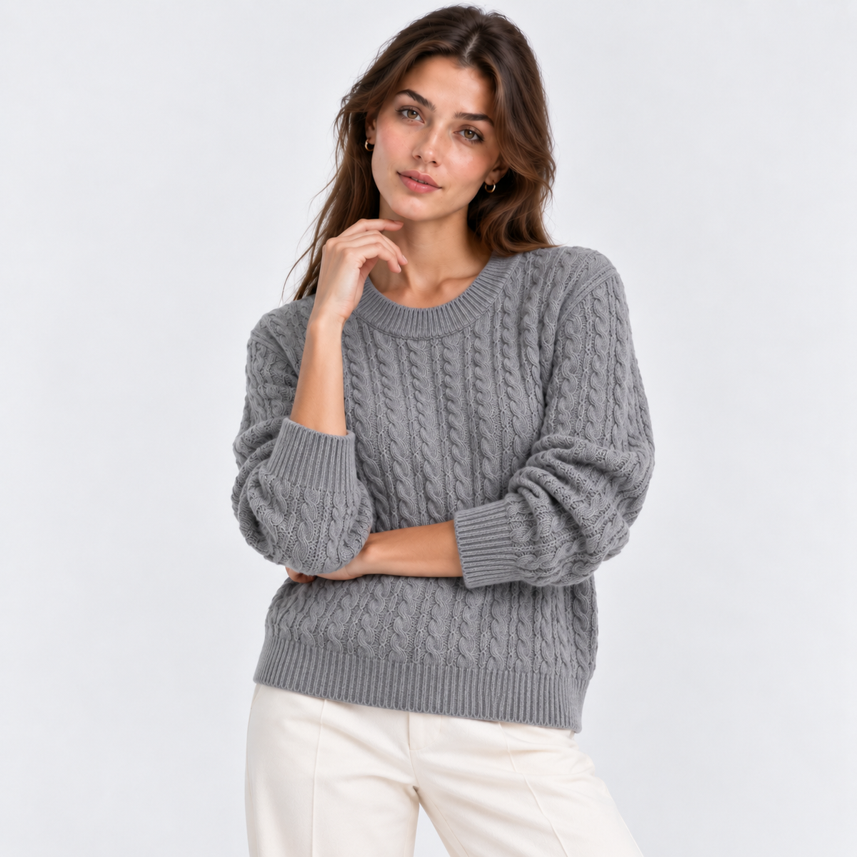 A woman wearing a gray cable-knit crewneck sweater and cream pants, posing against a light gray studio background.