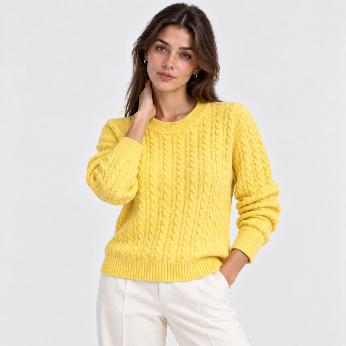A woman wearing a bright yellow cable-knit sweater and white trousers, posing against a light gray studio background.